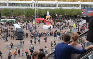 Menschen auf einer Rolltreppe schauen auf viele Menschen, die auf einem Platz an verschiedenen Ständen anstehen.