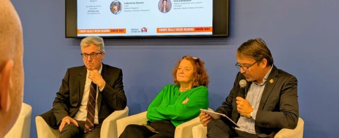 Zwei Männer und eine Frau sitzen vor einer blauen Wand. Einer der Männer spricht in ein Mikrofon.