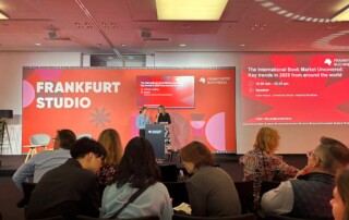 Ein Publikum sitzt vor einer Bühne, am Rednerpult stehen zwei Frauen.