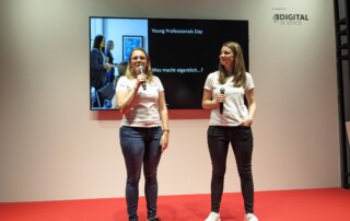 Zwei Frauen in weißen T-Shirts halten Mikrofone in den Händen und stehen auf einer Bühne mit rotem Boden.