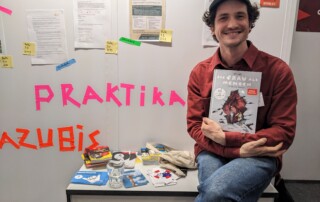 Ein Mann sitzt vor einer weißen Wand mit Aushängen und hält ein Buch in den Armen.