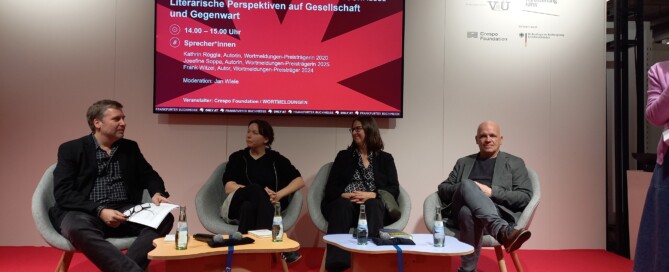 Zwei Männer und zwei Frauen sitzen auf grau bezogenen Stühlen in einer Reihe und sind im Gespräch. Hinter ihnen hängt ein Bildschirm, welcher den Veranstaltungstitel und die Namen der Teilnehmenden anzeigt. Die Bühne, auf der sie sich befinden, hat einen roten Boden.