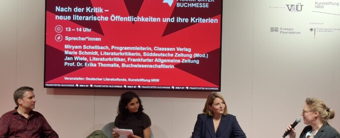 Drei Frauen und ein Mann sitzen auf grau bezogenen Stühlen auf einer Bühne mit rotem Boden und sprechen miteinander. Hinter ihnen hängt ein Bildschirm, welcher den Veranstaltungstitel und die Namen der Teilnehmenden anzeigt.
