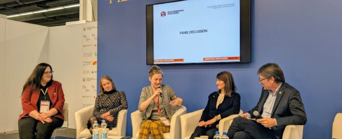 Vier Frauen und ein Mann sitzen vor einer blauen Wand. Eine Frau spricht in ein Mikrofon, die anderen hören ihr zu.