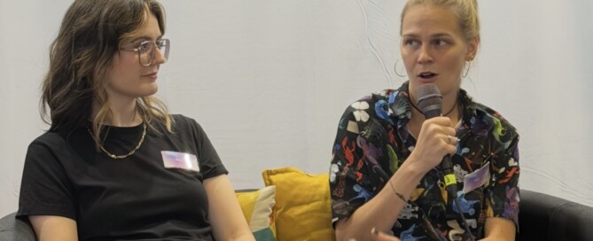 Zwei Frauen sitzen auf einem Sofa, eine von ihnen spricht in ein Mikrofon.