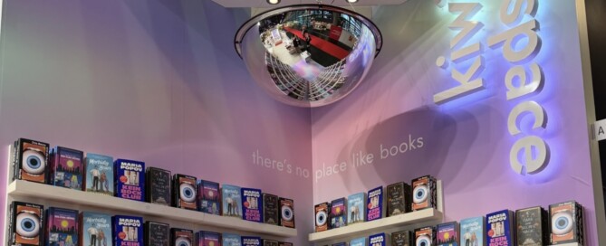 Der Ausstellungsraum ist mit Büchern in Regalen und einer spiegelnden Halbkugel an der Decke ausgestattet. An der Wand steht „There's no place like books” und es hängt eine beleuchtete Aufschrift mit der Aufschrift „kiwispace”.